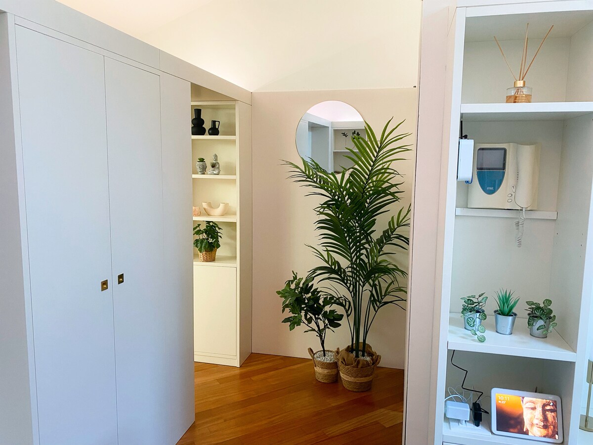 An open space showcases modern cabinetry in light tones. A rounded mirror enhances the room's depth. Potted plants introduce greenery, positioned elegantly near the wall. A shelf displays decorative items and additional plants, contributing to a bright and inviting atmosphere.