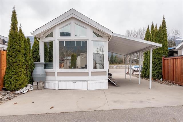 A modern RV structure features large windows, allowing natural light to fill the interior. The roof overhang provides shade, while neatly trimmed evergreen trees flank the entrance. A paved driveway leads to a covered parking area, enhancing accessibility.