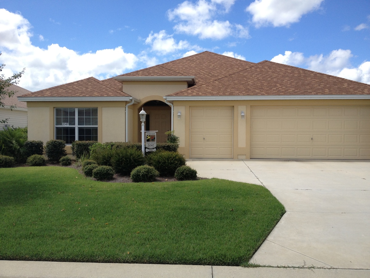A single-story home with a warm yellow exterior features a brown tiled roof and a two-car garage. Lush green landscaping surrounds the entryway, with manicured bushes and a neatly trimmed lawn. Large windows allow natural light into the inviting living space.