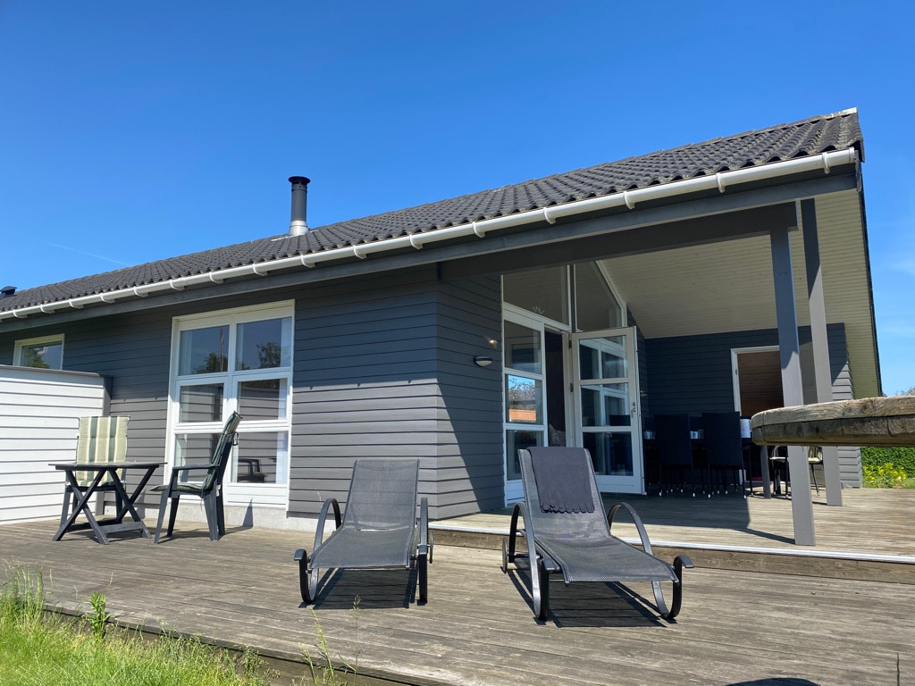 A single-story house is shown with a blue-grey exterior and a spacious wooden deck. Two reclining chairs are positioned on the deck, accompanied by an outdoor dining set in the background. Large windows provide ample natural light, contributing to an open and inviting atmosphere.