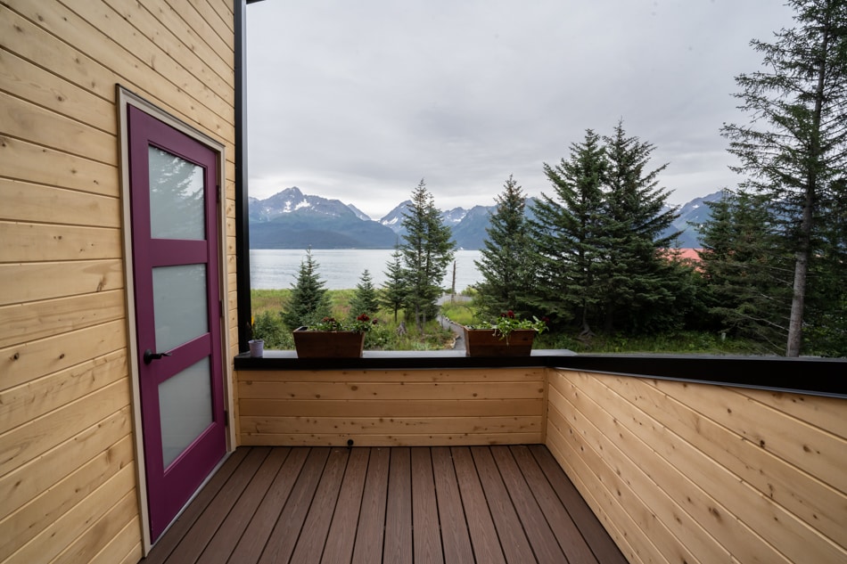 A deck area is presented with wooden flooring, bordered by a smooth wooden railing. Flower pots are positioned along the edge, while views of mountains and Resurrection Bay are visible in the background, framed by evergreen trees.