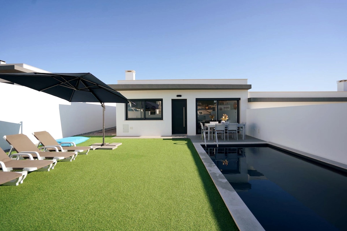 An outdoor area features a sleek pool framed by artificial grass, with four loungers positioned nearby. A shaded dining table is set for meals, and an umbrella provides additional cover from the sun. The house's contemporary design is visible through large glass doors.