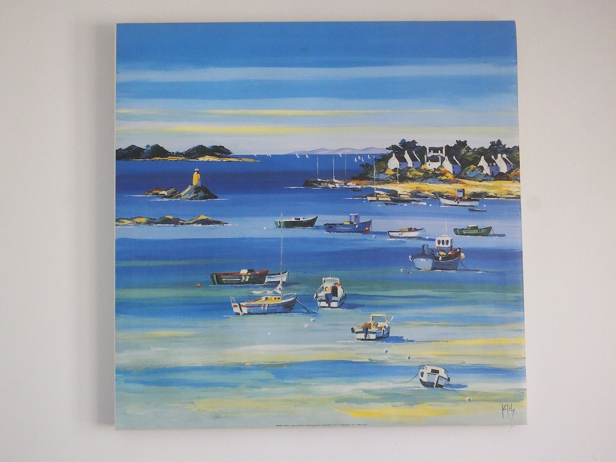 A vibrant painting depicts a serene coastal scene with boats gently floating on clear blue waters. The shoreline is lined with greenery and buildings, while distant islands are visible under a bright, cloudless sky.