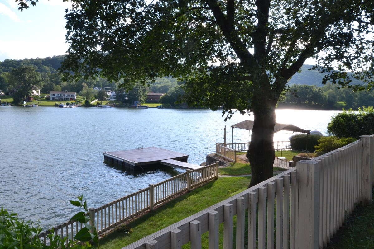 A view of a serene lake is framed by a large tree, with a private dock extending into the water. The grassy area leads to the dock, and a covered seating area can be seen beside the water, offering a peaceful spot to relax.