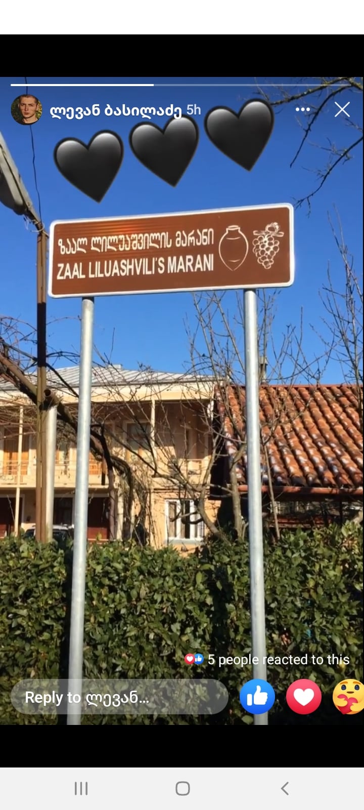 A brown sign is displayed in front of a traditional building, indicating 'Zaal Lluashvili's Maran.' The background features a rustic structure with a red-tiled roof. Sunlight illuminates the area, enhancing visibility of the sign and surroundings.