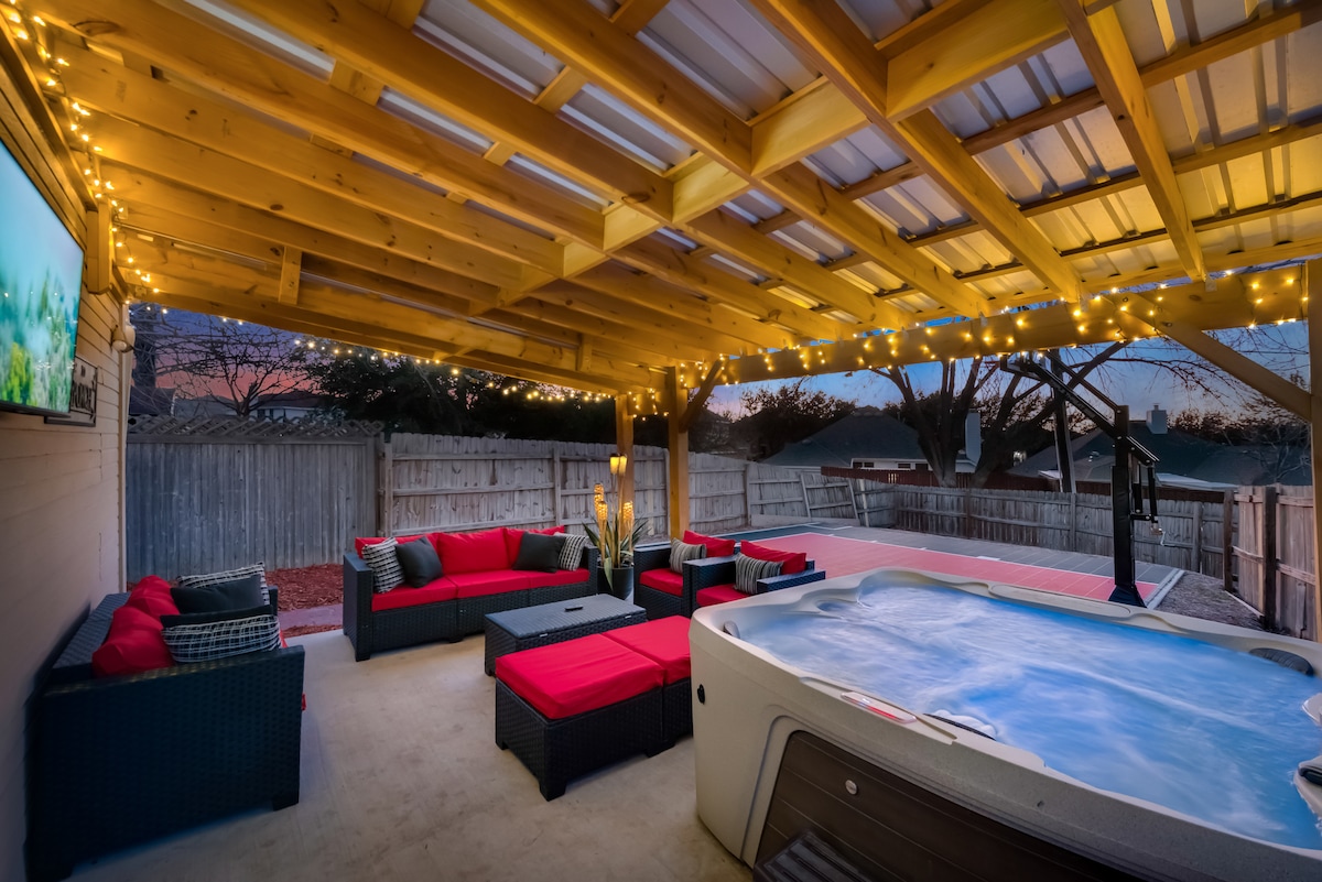 An outdoor lounge area is sheltered beneath a wooden pergola, illuminated by string lights. Cozy seating with bold red cushions surrounds a hot tub, offering a relaxed atmosphere for gatherings. A flat-screen TV is mounted on the wall, providing entertainment options.