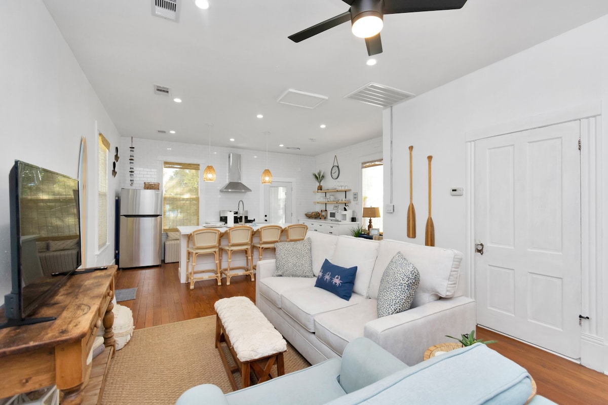 An open living space includes a light-colored sectional sofa and a wooden coffee table, oriented towards a television. A dining area with four wicker chairs is adjacent to a kitchen featuring modern appliances. Natural light filters through the windows, creating a bright ambiance.