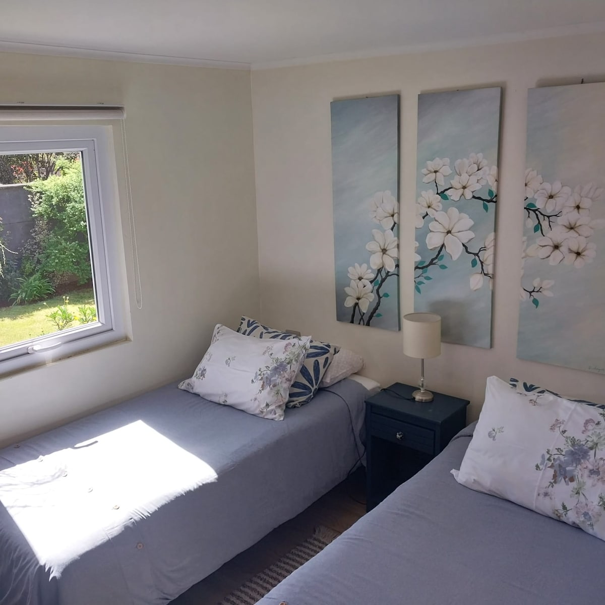 A bright bedroom features two beds dressed in muted linens, complemented by decorative pillows. A nightstand with a lamp sits between the beds. Natural light enters through a window, illuminating floral artwork that enhances the calming ambiance.