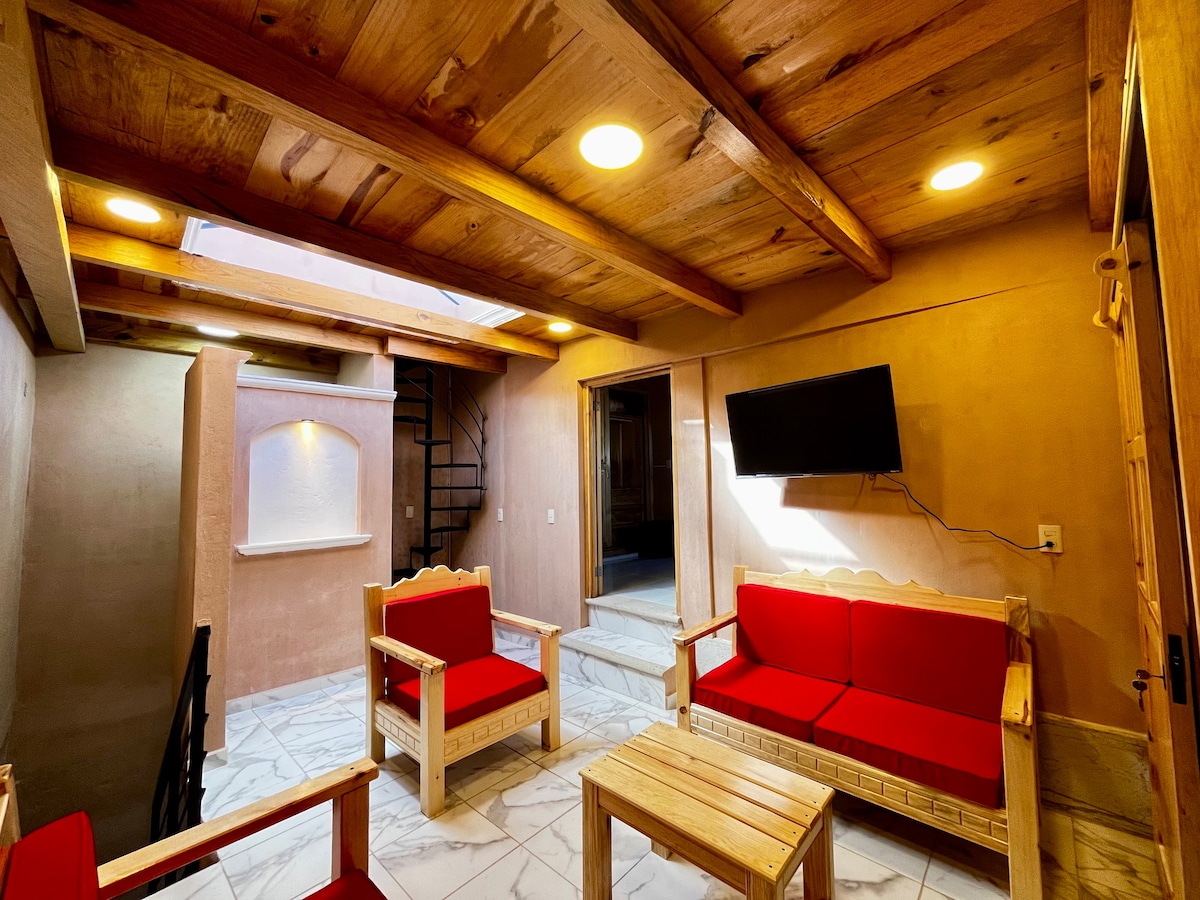 A cozy lounge area is presented, featuring two red sofas and two wooden chairs arranged around a small wooden table. Natural light enters through a skylight, illuminating the rustic wooden beams overhead and enhancing the warm tones of the plastered walls.