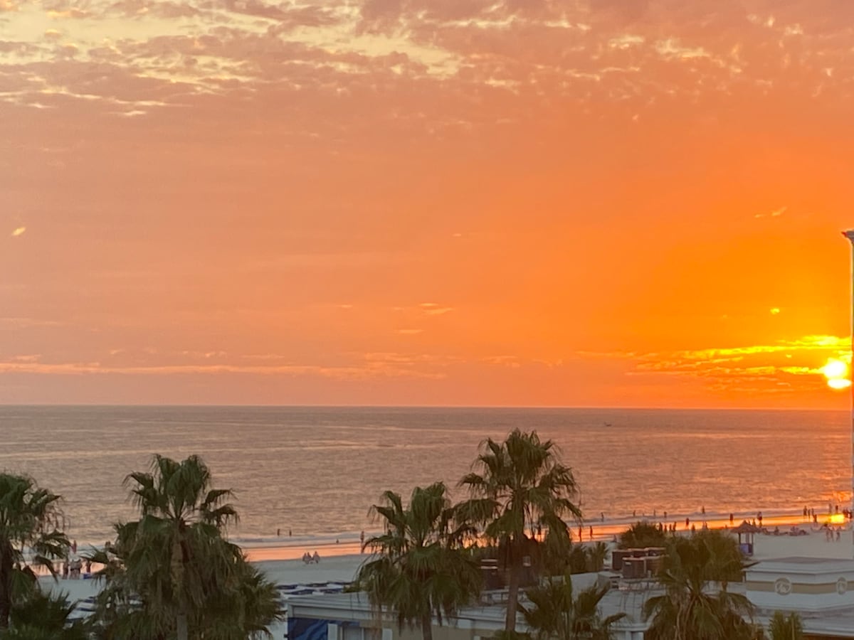 A vibrant sunset casts warm orange and yellow hues across the sky, with the sun nearing the horizon over the calm ocean. Silhouettes of palm trees frame the scene, while people stroll along the beach, creating a serene coastal atmosphere.