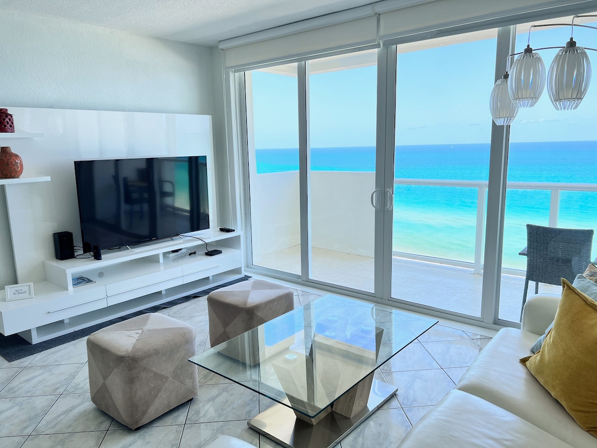 A spacious living area features a modern glass coffee table and a sofa, complemented by decorative pillows. Large sliding glass doors offer unobstructed ocean views, while a flat-screen TV is placed on a sleek white media console. A dining chair is visible in the corner.