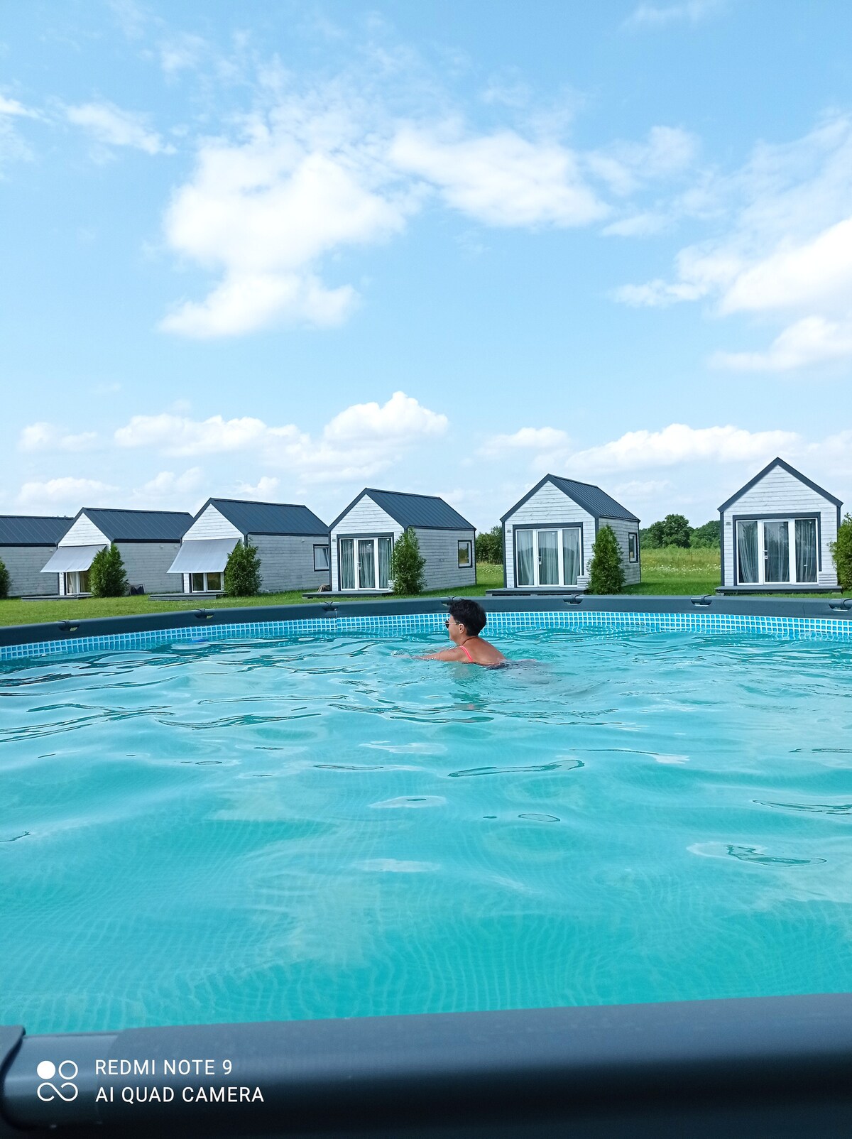 A swimming pool is seen in the foreground, with a person enjoying the water. In the background, several modern cabins are arranged on a grassy area under a clear blue sky, adding to the serene environment.