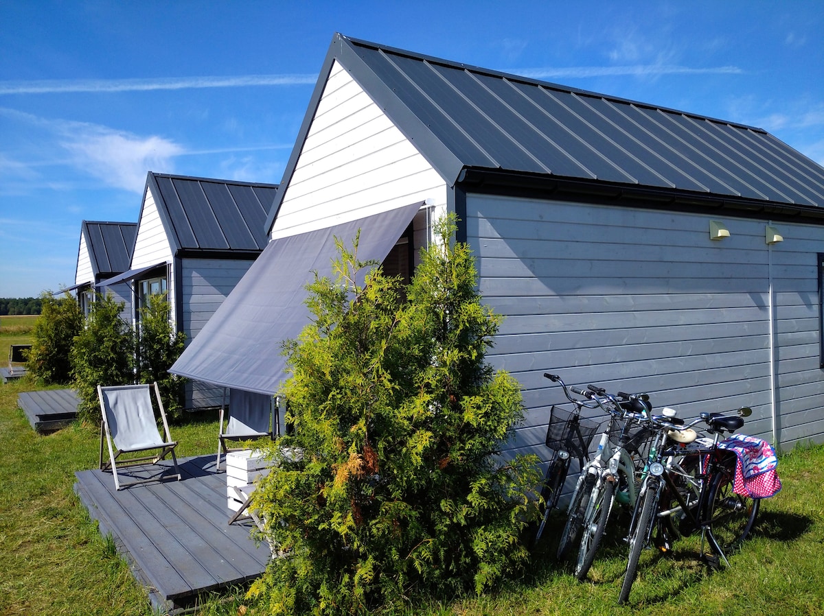 Modern Scandinavian-style cabins are arranged in a row, showcasing a sleek design under a clear blue sky. Each cabin features a shaded outdoor area with seating. Bicycles are parked nearby, inviting exploration of the surrounding natural landscape.