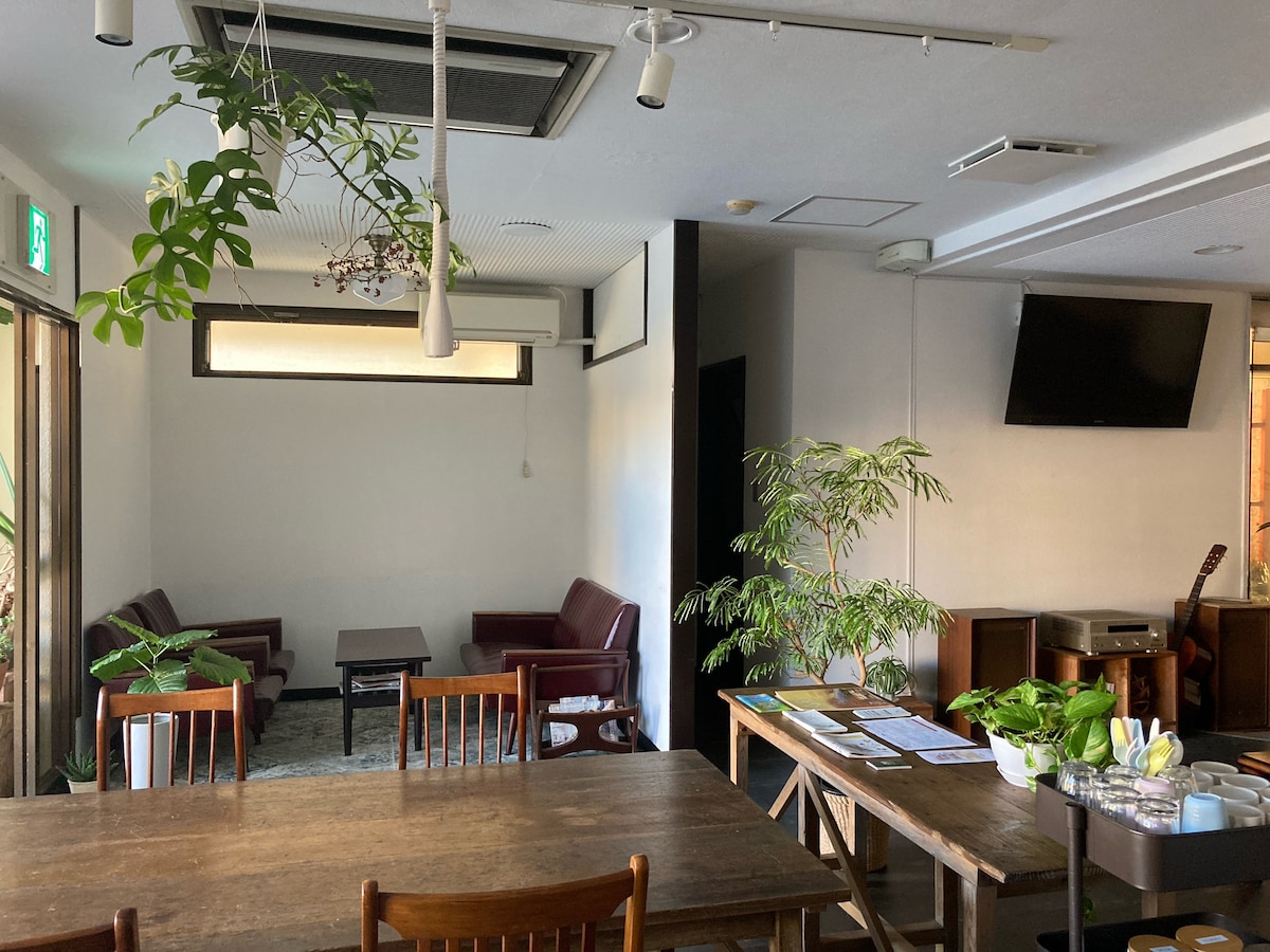 A shared lounge area features two burgundy sofas and a wooden coffee table at its center. Natural light enters through large windows, illuminating the greenery from nearby plants. A wooden dining table is visible in the foreground, complemented by a small display of potted plants.