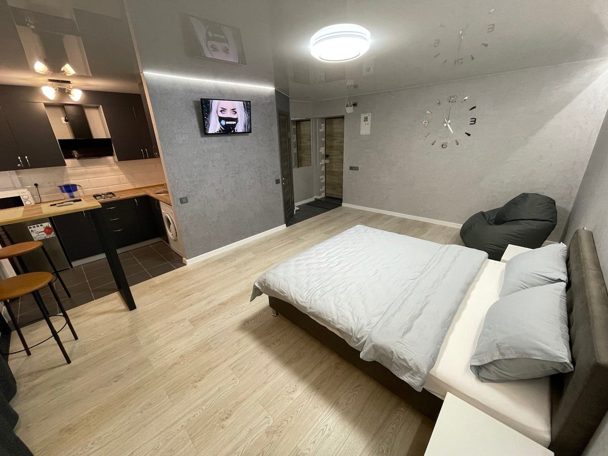 A minimalist studio apartment features a comfortable bed with grey bedding, a mounted TV, and a dining area with bar stools. A modern kitchen is visible in the background, with sleek cabinetry and a light-colored countertop. Natural light enters through a nearby window.