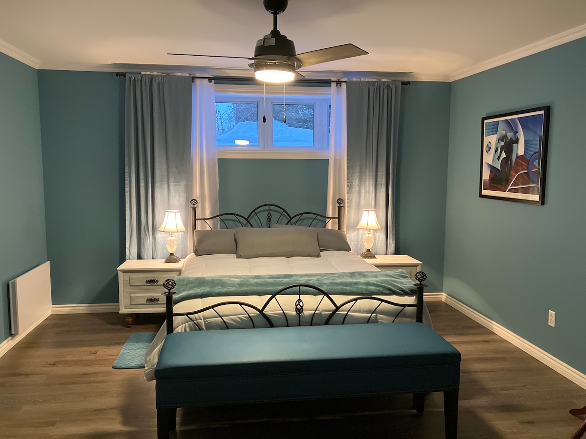 The spacious bedroom features a queen-sized bed with soft linens, flanked by two bedside tables with lamps. A blue bench is positioned at the foot of the bed. Light blue walls and curtains complement the decor, while a ceiling fan provides airflow.