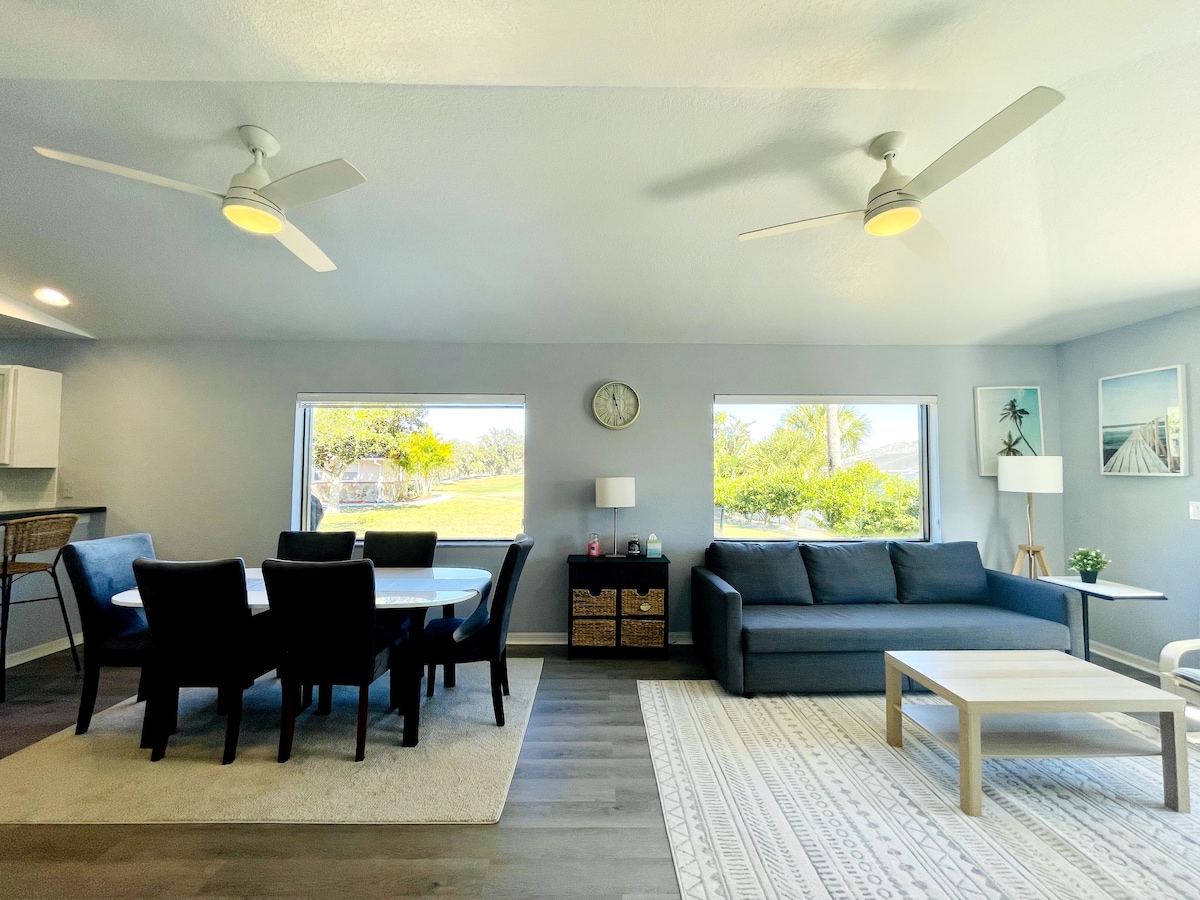 A spacious living and dining area features a dining table with black chairs and a sleek sofa positioned near large windows. Soft light enters through the windows, illuminating the light gray walls and wooden flooring. Ceiling fans contribute to the comfortable ambiance.