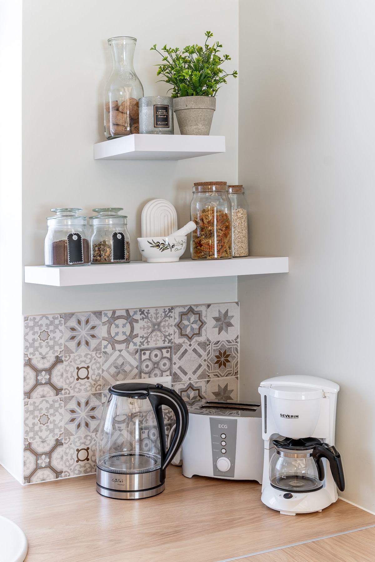 A well-organized kitchenette area features glass jars filled with various ingredients on floating shelves above a decorative tiled wall. A coffee maker and kettle stand on the countertop, alongside a compact coffee grinder, providing essential amenities for beverage preparation.