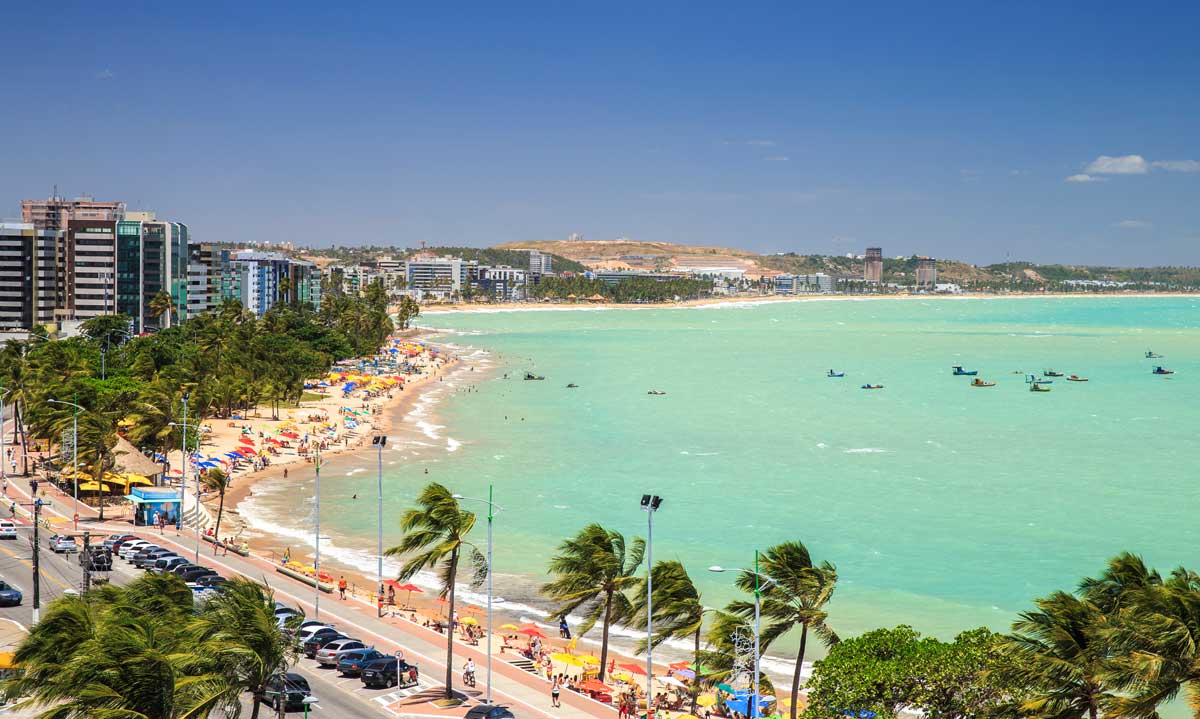 The image captures a lively beach scene lined with palm trees and sun umbrellas. Waves gently lap against the shore, while people are seen enjoying the sandy beach and water activities. Tall buildings are visible in the background, providing a contrast to the natural scenery.