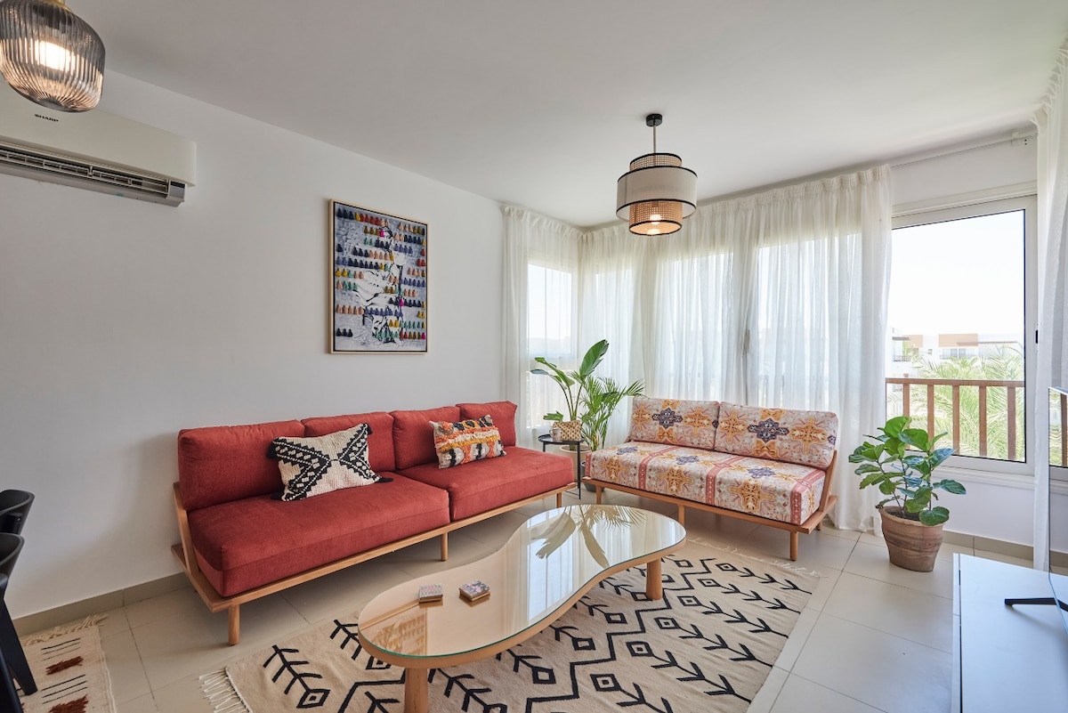 A bright living space features a coral-colored sofa and a patterned loveseat, complemented by a glass coffee table. Natural light fills the room through large windows, with sheer white curtains softly filtering the sunlight. A green potted plant adds a touch of nature, and artwork adorns the wall.