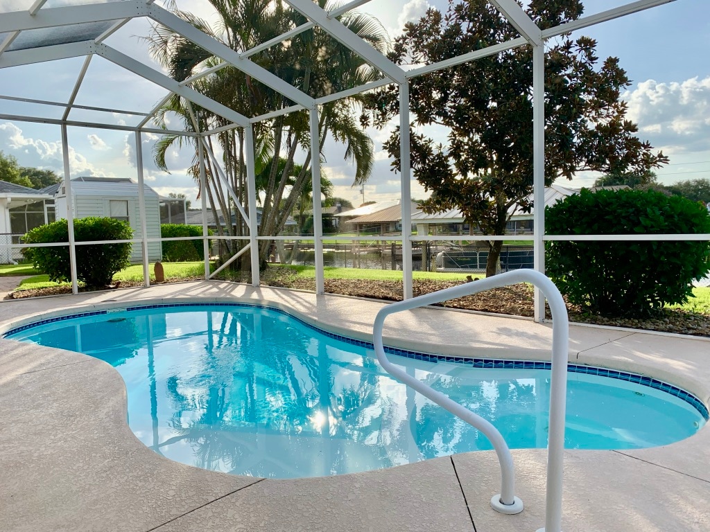 A screened-in pool area features a refreshing, clear pool surrounded by lush greenery. The pool's gentle curves are framed by a sturdy white railing, while a variety of palm trees can be seen beyond the enclosure. Natural light brightens the space.