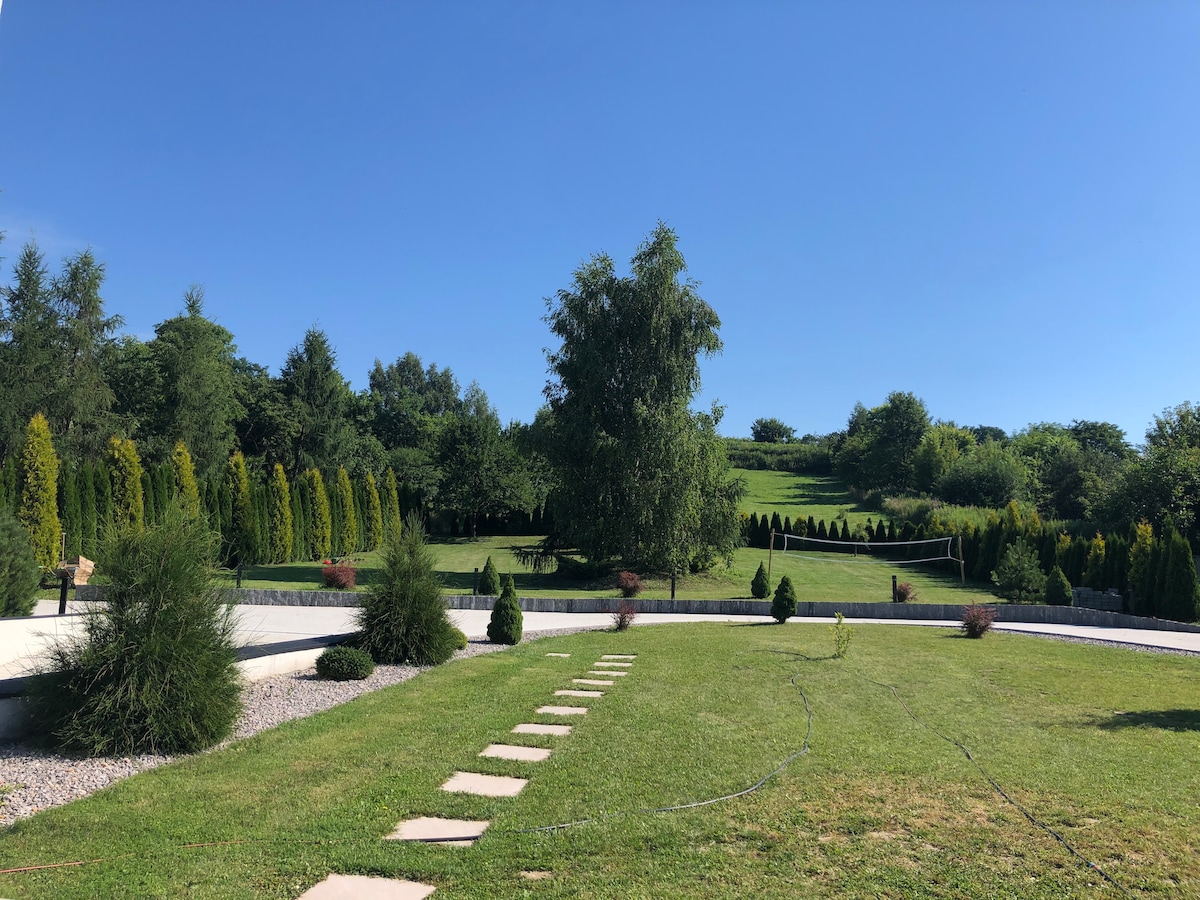 A well-maintained garden is presented, featuring a lush lawn bordered by neatly trimmed shrubs and trees. Stone pathways lead through the greenery toward the surrounding nature, creating an inviting outdoor space ideal for relaxation and play.