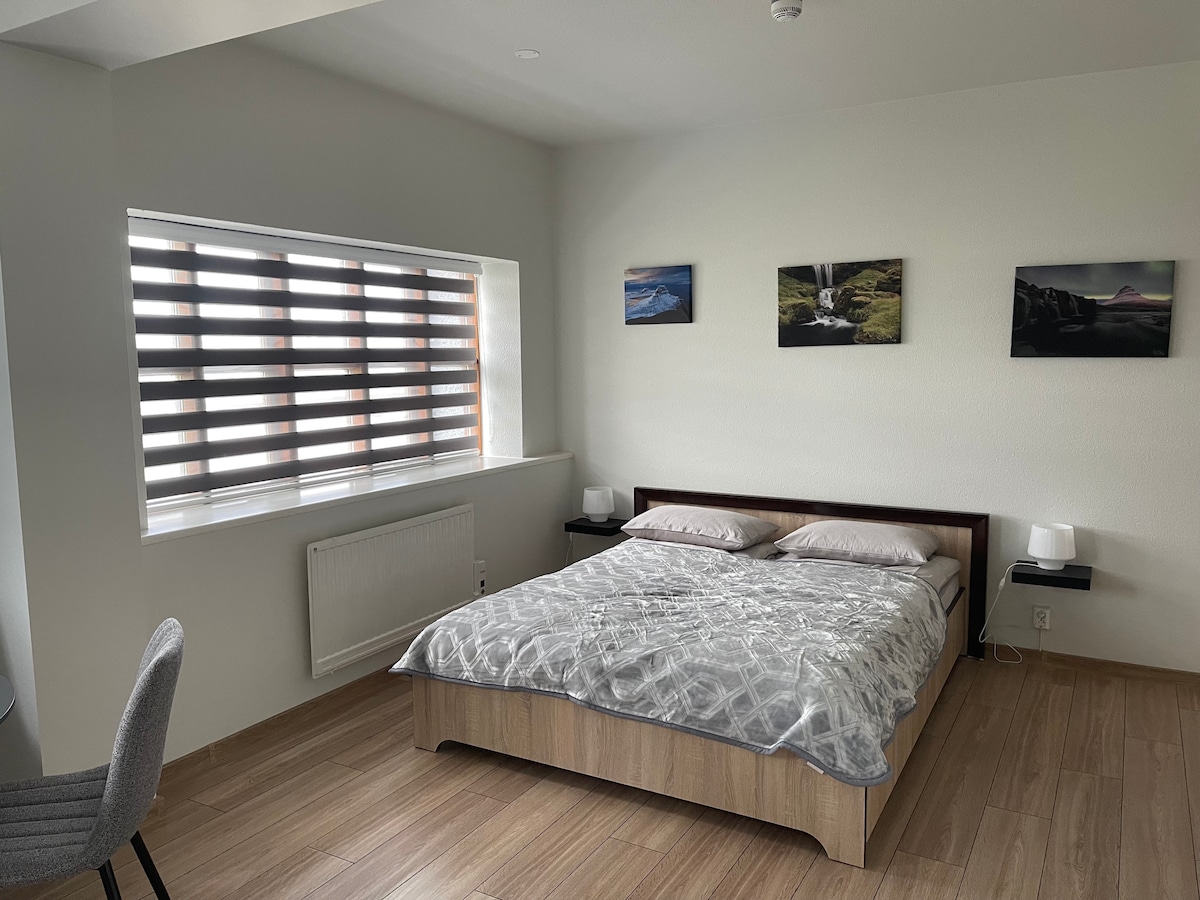 The bedroom features a spacious queen-sized bed with a light-colored bedspread. Two bedside tables with lamps are positioned on either side. Above the bed, three framed photographs add visual interest. A window with horizontal slats provides natural light while maintaining privacy.