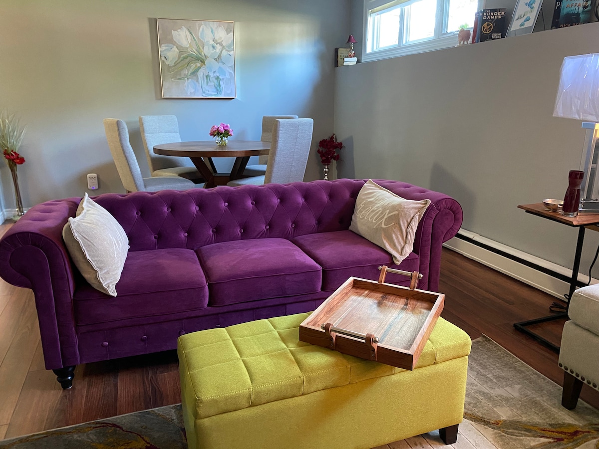 A vibrant purple tufted sofa is positioned in the living area, accompanied by two decorative pillows. A yellow ottoman with a wooden tray sits in front. A round dining table with white chairs is visible in the background, complemented by floral decor in the corner.