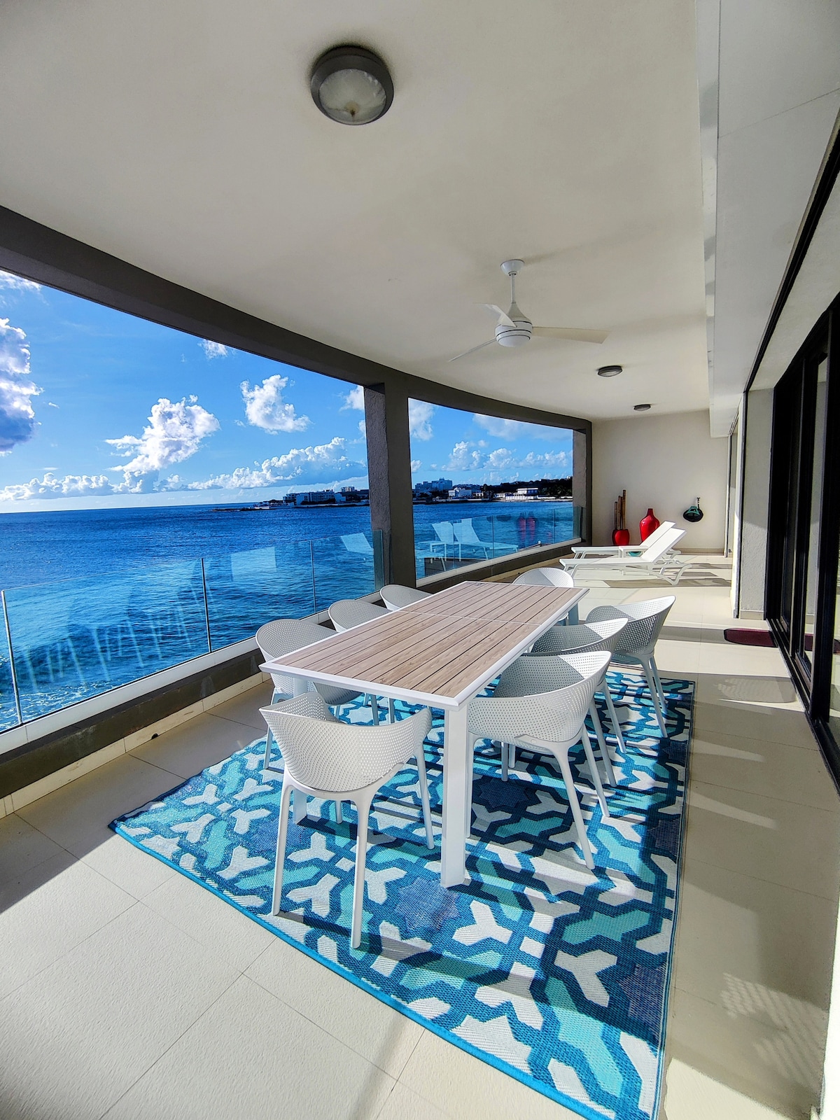An outdoor dining area is seen on a spacious terrace, furnished with a white table and six matching chairs. A vibrant blue patterned rug adds color underfoot. The backdrop features panoramic views of the Caribbean Sea, complemented by bright blue skies and fluffy clouds.