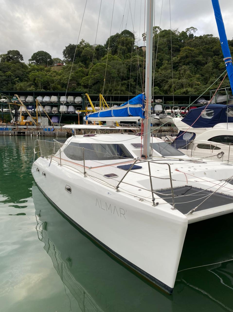 A sleek catamaran is docked at a marina, featuring a spacious deck and modern lines. Lush greenery is visible in the background, with numerous boats stored in the facility. The calm water reflects the vessel, indicating a serene environment.