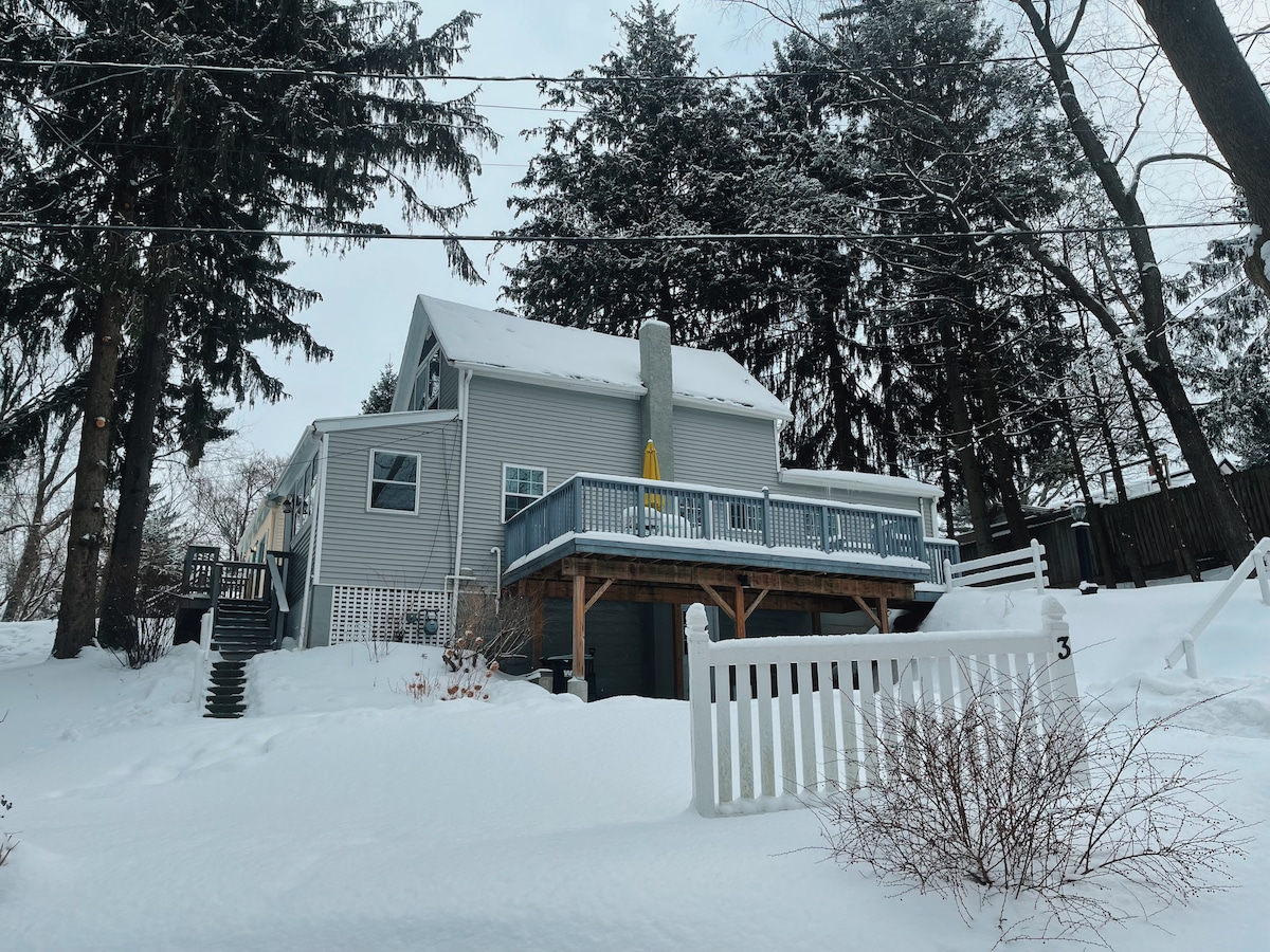 The exterior of a two-story cottage features a large deck with a yellow kayak resting against the wall. Snow blankets the ground, and tall trees provide a natural backdrop. A white picket fence defines the front yard, enhancing the inviting atmosphere of the property.