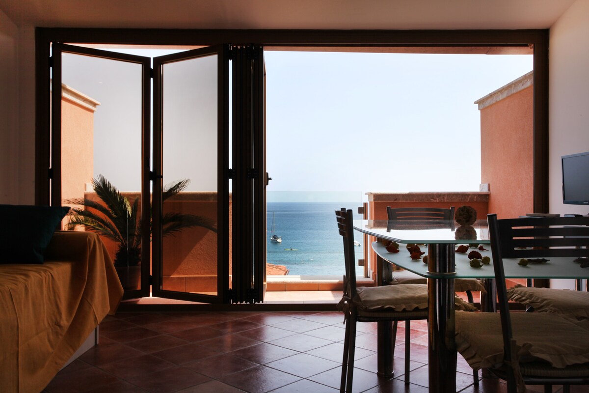 A spacious living area features large open glass doors, providing an unobstructed view of the sea. A dining table with light-colored chairs sits in the foreground, with a sofa in soft hues positioned next to it. Natural light floods the room.