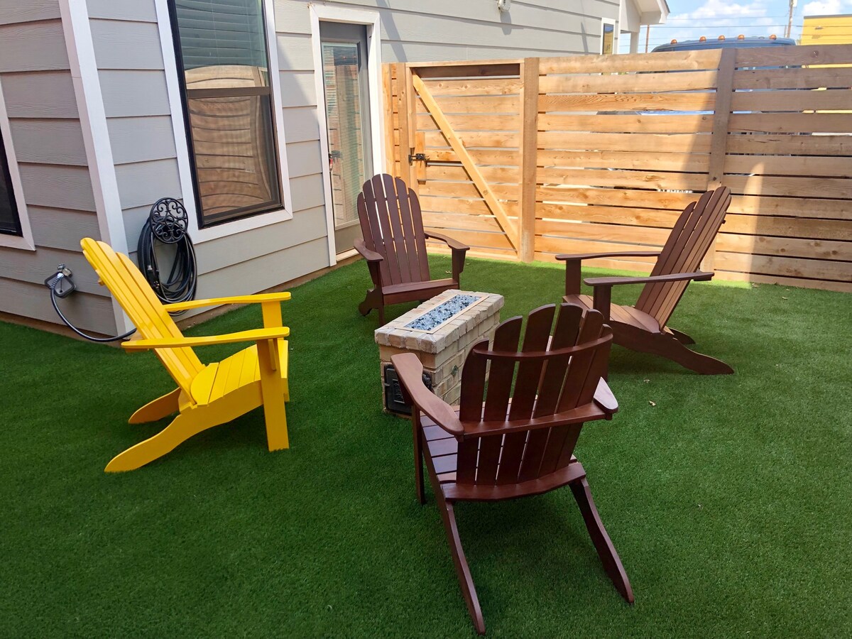 A private outdoor area features four Adirondack chairs arranged around a stone fire pit, situated on well-maintained artificial grass. A wooden fence provides privacy, while a wall-mounted charging station for electric vehicles is visible nearby.