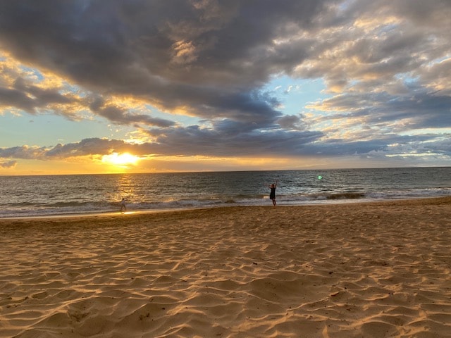 A sandy beach is depicted at sunset, with warm hues reflecting off the water. Gentle waves lap at the shore. Silhouettes of individuals can be seen walking along the water's edge against a backdrop of colorful clouds.