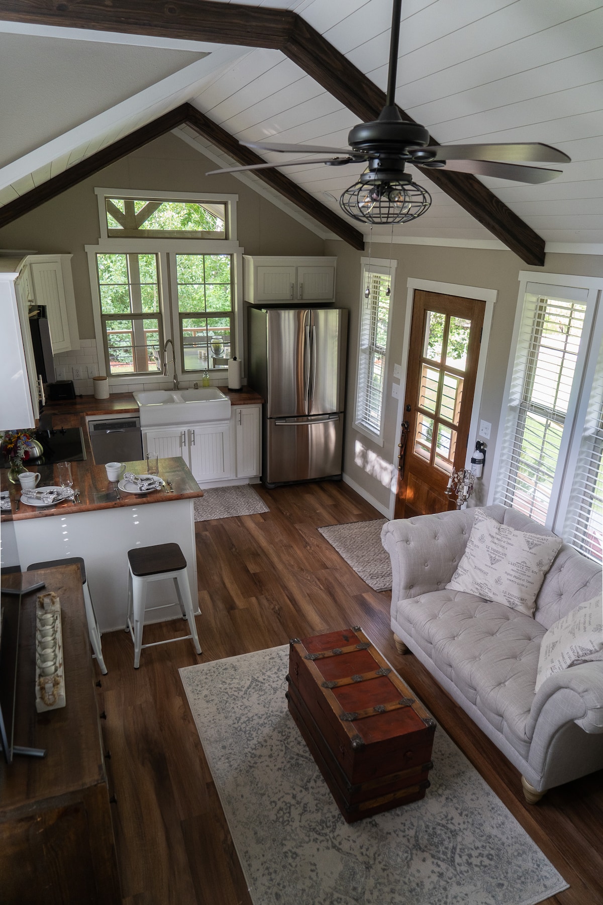 An open living area features a plush sofa positioned near a wooden coffee table and a rug. A well-equipped kitchen is visible in the background, complete with a stainless steel refrigerator and counter seating. Natural light streams through multiple windows, enhancing the inviting space.