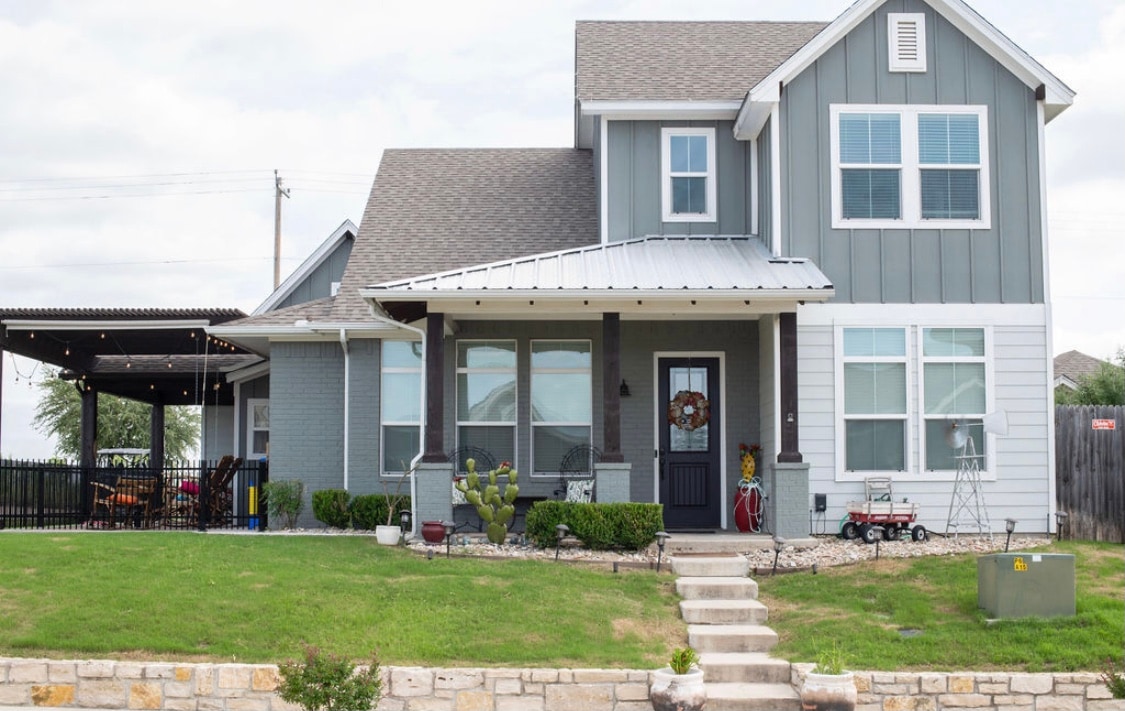 A two-story house is presented with a gabled roof and a welcoming front porch. Lush green grass and gravel pathways enhance the landscape. Large windows offer abundant natural light, while a covered patio area features outdoor furniture, perfect for relaxation. A small fenced area is visible on one side.