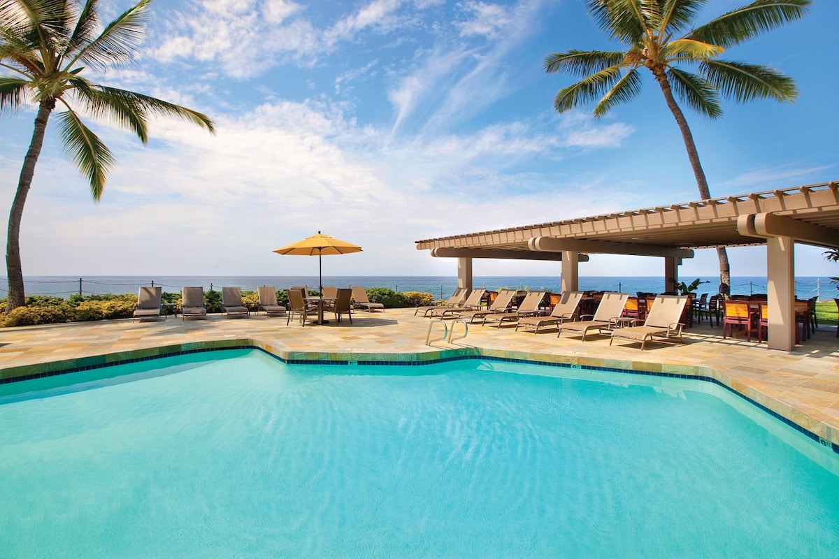 A shimmering pool area is surrounded by lounge chairs under a clear blue sky. Tropical palm trees sway gently nearby, and a large umbrella provides shade. The ocean is visible in the background, enhancing the relaxing atmosphere of the outdoor space.