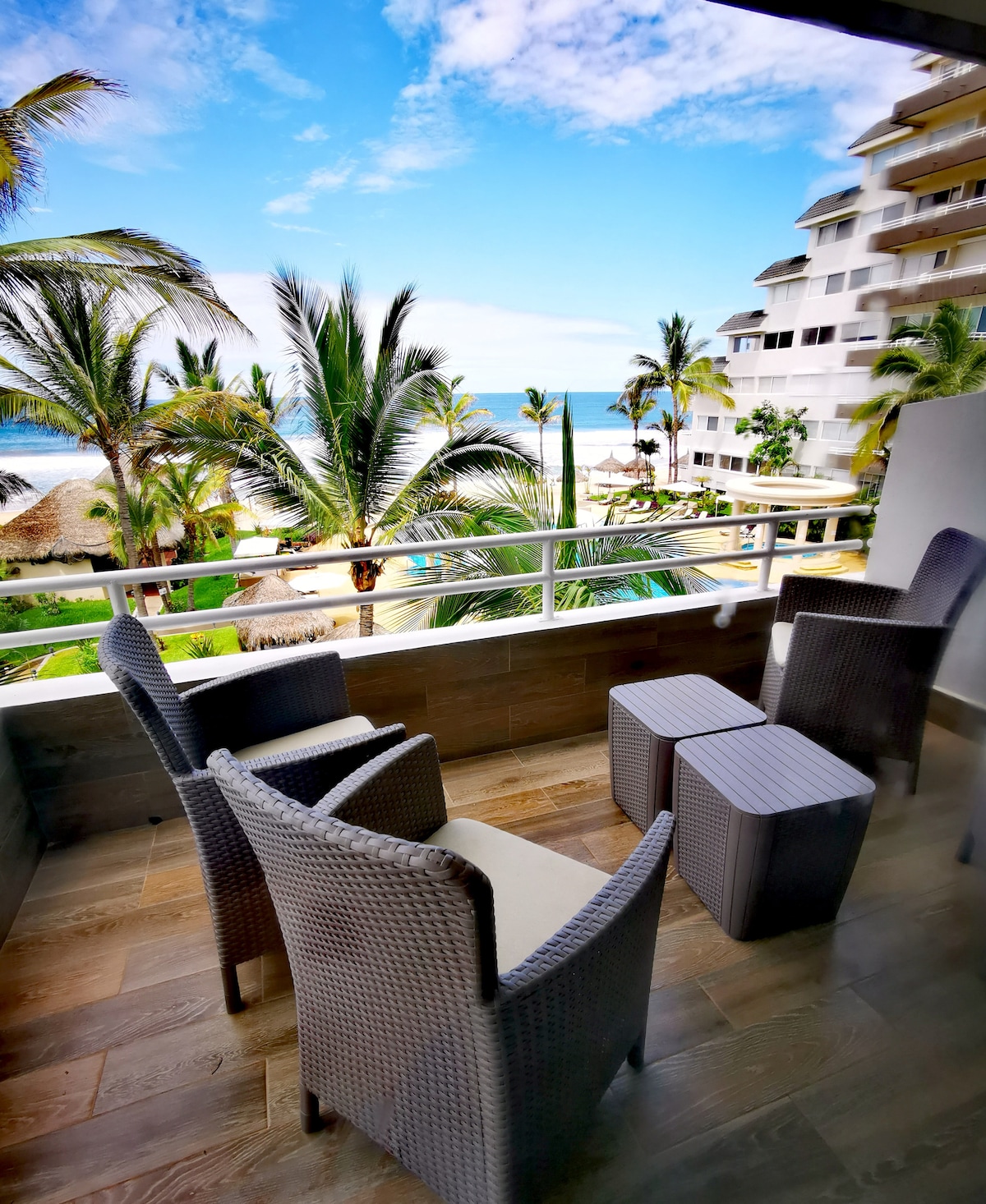 A set of two comfortable chairs and a small table occupies a balcony overlooking lush palm trees and the beach. The sea can be seen in the distance, with a clear sky creating a serene atmosphere. The space is designed for relaxation.