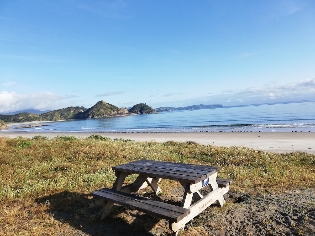 A sturdy wooden picnic table sits on a grassy area overlooking a calm beach. The serene waters reflect a clear sky and distant hills, creating a peaceful coastal scene perfect for outdoor dining or relaxation.