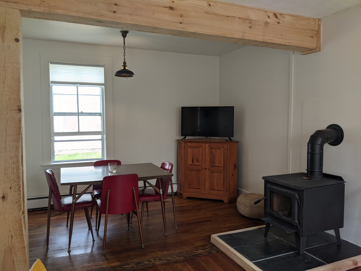 A cozy living area features a wood stove and a television across from a dining table surrounded by red chairs. Light enters through a window, enhancing the warm wood flooring and inviting atmosphere.