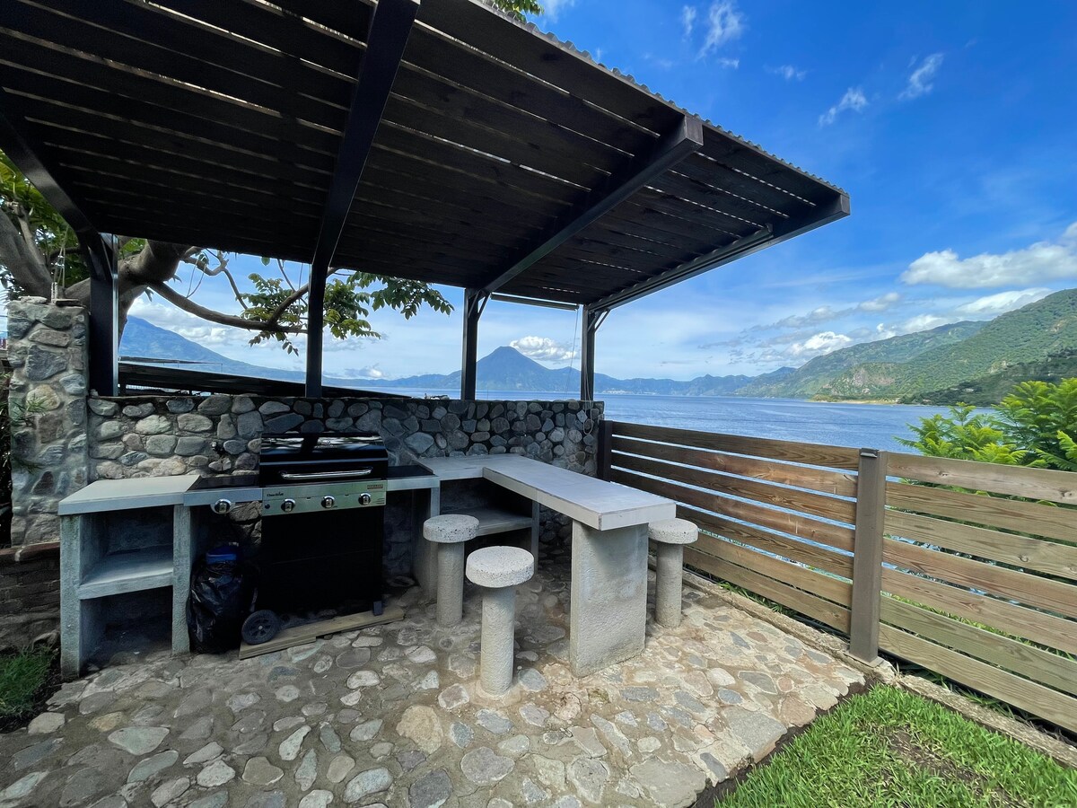 An outdoor cooking area features a covered grilling space with a stone wall backdrop. The setting includes a stone table and seating, offering views of the lake and surrounding mountains. Natural light brightens the area, complemented by a clear blue sky.