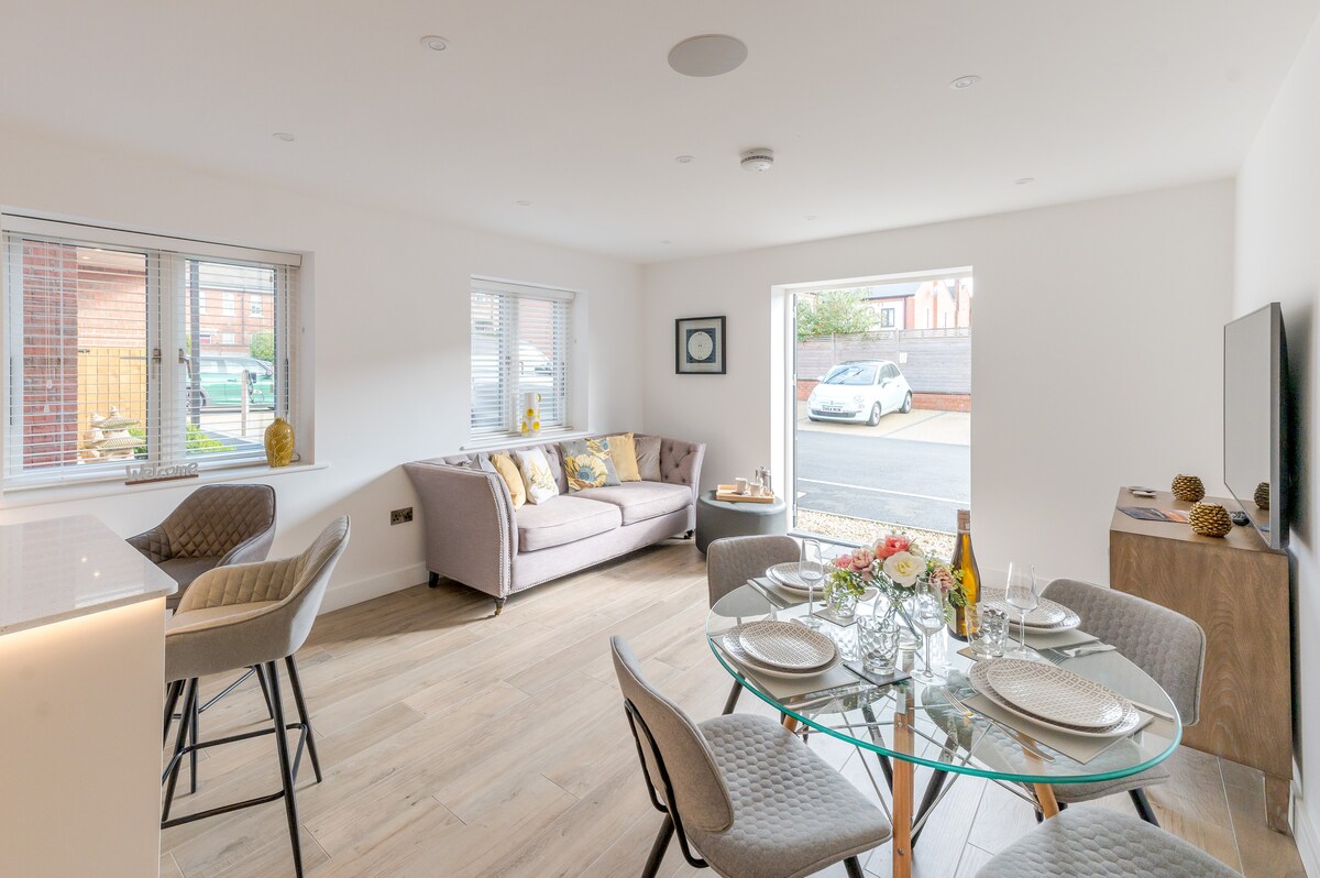 An open-plan living area features a modern kitchen with a dining table set for four, and a cozy seating area with a neutral-toned sofa. Large windows provide natural light and a view of the exterior. The space presents a contemporary design with clean lines and a welcoming ambiance.