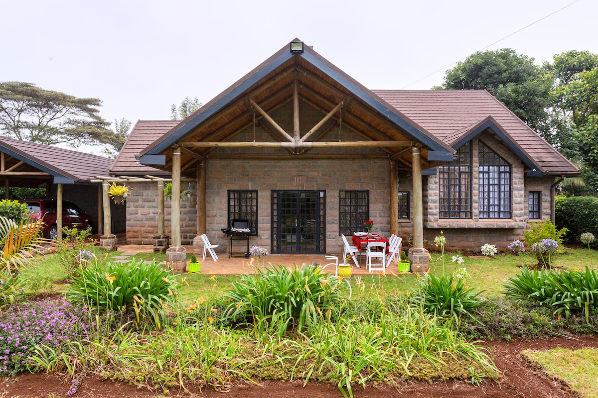 A single-story stone house is set in a lush garden featuring various plants and flowers. A covered porch with white chairs and a small table provides outdoor seating, while large windows allow for natural light to fill the interior.