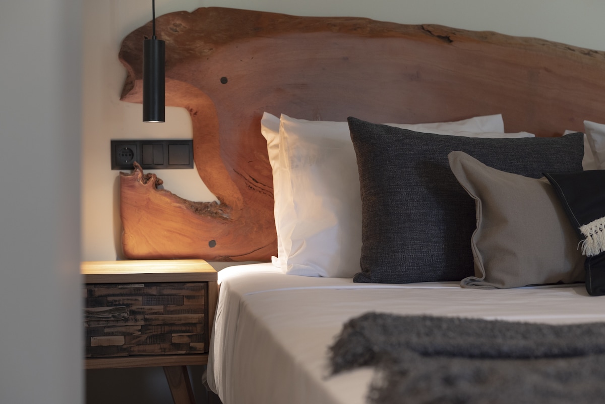 A spacious bed is positioned against a striking wooden headboard, crafted from natural materials. Soft, neutral-toned pillows are layered atop the white linens. A bedside table, with an intricate wooden design, is accompanied by a sleek pendant light, adding subtle illumination.