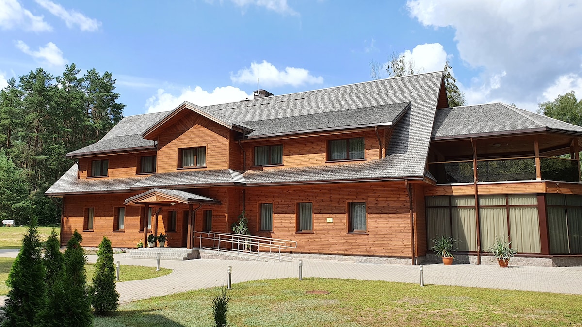 A wooden building with a sloped roof and a welcoming entrance is set amidst greenery. Large windows allow natural light into the interior, while the surrounding landscape features well-maintained grass and neatly arranged shrubs.