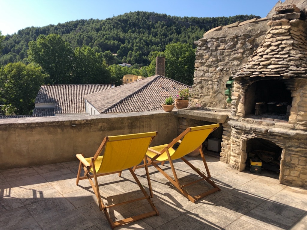 A terrace features two wooden lounge chairs with yellow fabric, situated on a stone surface. In the background, a view of lush green hills is visible, framed by historic stone buildings with terracotta roofs.