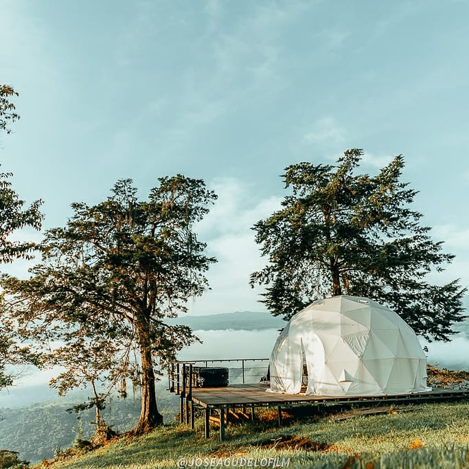 A geodesic dome is set on a wooden platform amidst lush greenery, with towering trees on either side. The serene landscape features rolling hills and a cloudy sky, creating a peaceful atmosphere for an outdoor escape.