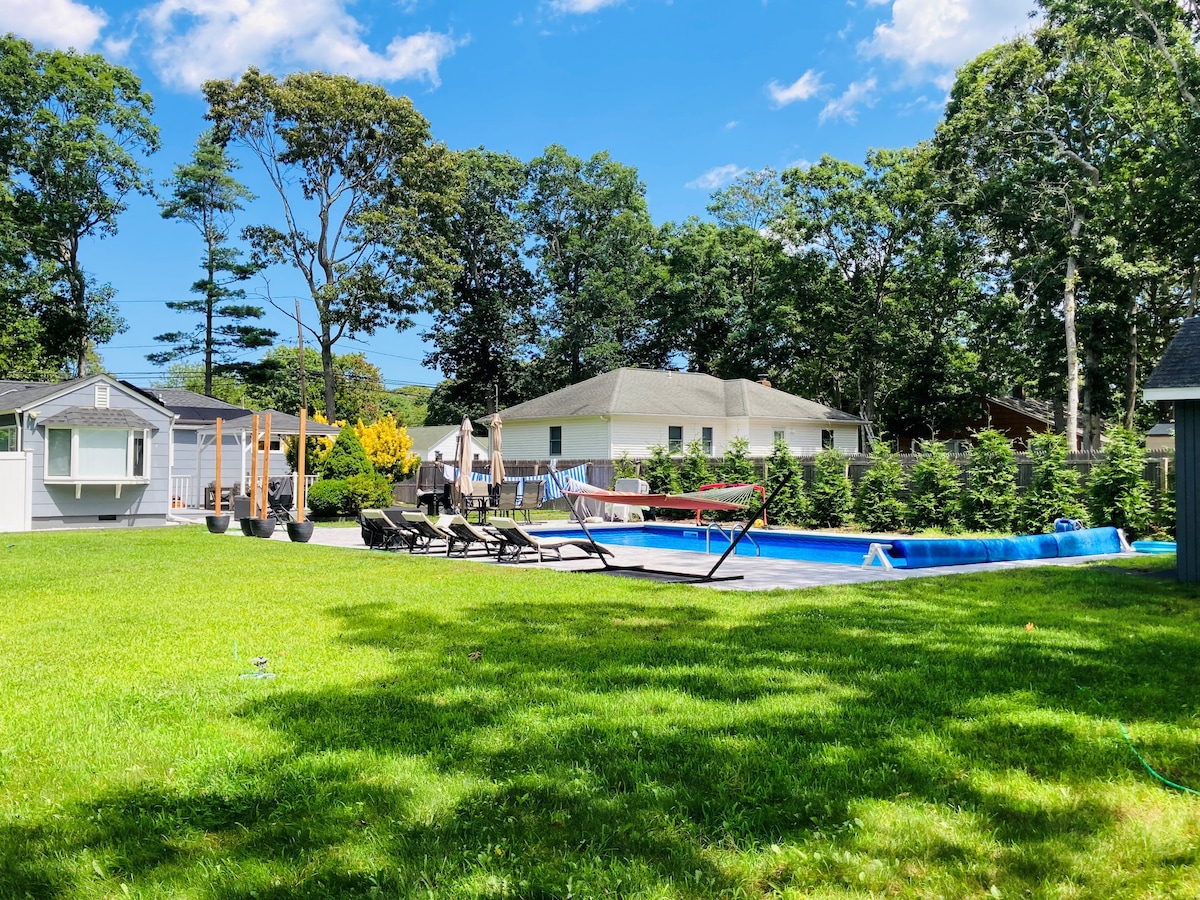 A spacious outdoor area is featured, highlighted by a heated pool surrounded by lounge chairs. Green grass extends throughout the yard, with trees providing shade in the background. A pergola is visible, creating an inviting space for outdoor gatherings.