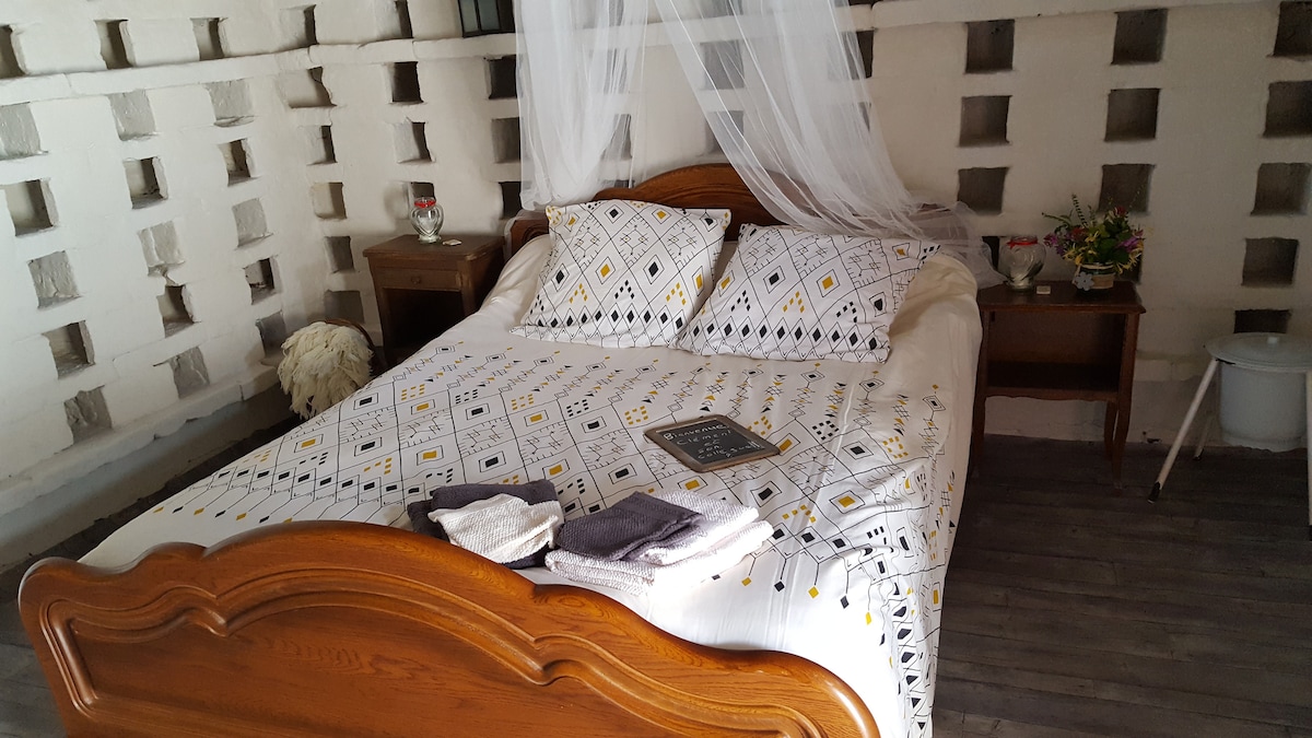 A spacious bed is centered in the room, featuring a decorative cover and two patterned pillows. Soft towels are neatly arranged at the foot of the bed. Natural light filters through the walls, which are accented with square openings, enhancing the rustic charm.