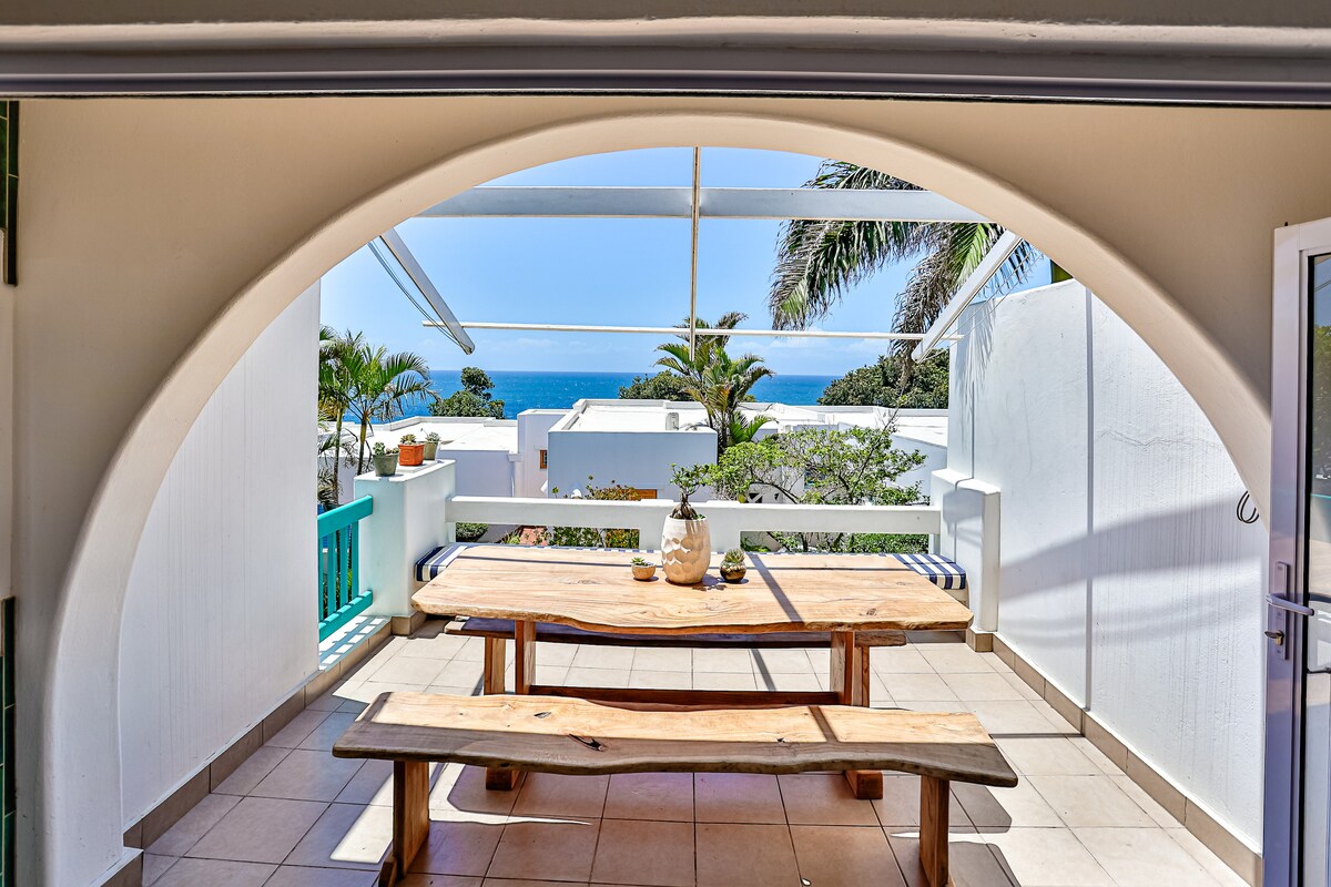 A spacious outdoor dining area is highlighted by a large wooden table with benches, set under a covered terrace. The backdrop features views of the ocean and palm trees, with natural light streaming in through an arched opening above.